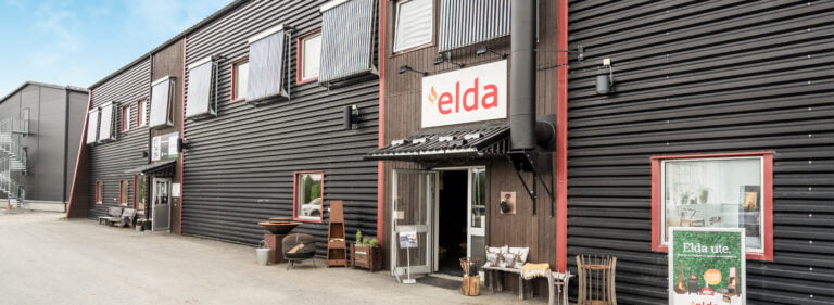 Svart byggnad med elda-skylt, entré och reklam för utomhusprodukter. Trädgårdsmöbler och dekor utanför på grusväg. Klarblå himmel i bakgrunden.