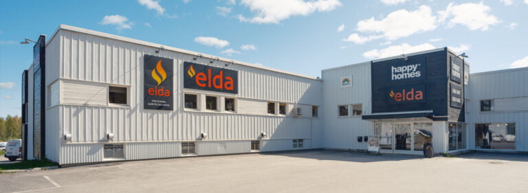 Vit byggnad med logotyper för Elda och Happy Homes på fasaden under en klarblå himmel. Asfalterad parkeringsplats framför byggnaden.