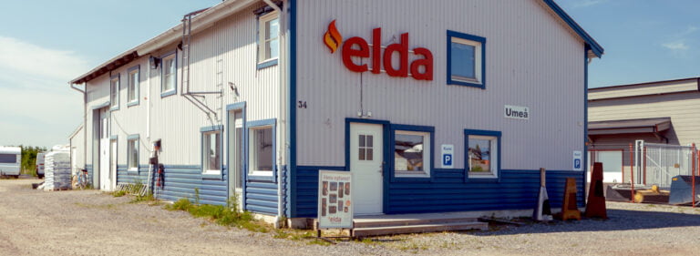 Butiksfasad med Elda-logotypen i rött på en vit och blå byggnad i Umeå. Kundparkering finns framför butiken.