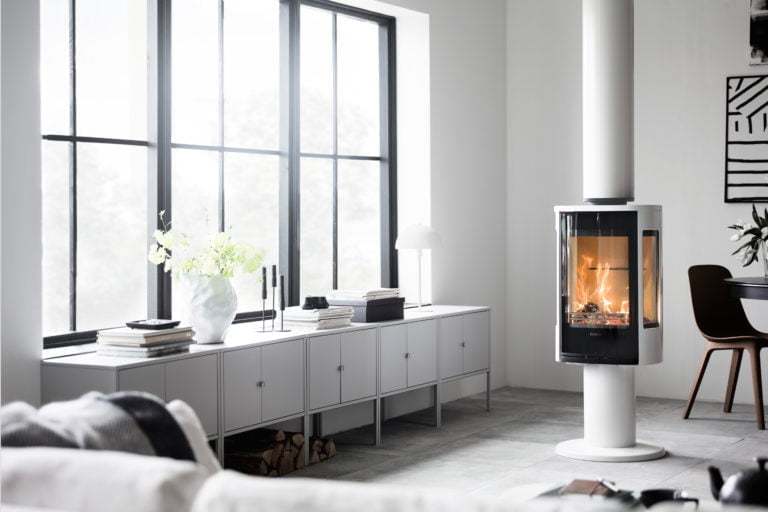 Modern inredning med eldstad, stora fönster och stilren möbel med blommor och böcker. Elden skapar en varm atmosfär i det ljusa rummet.