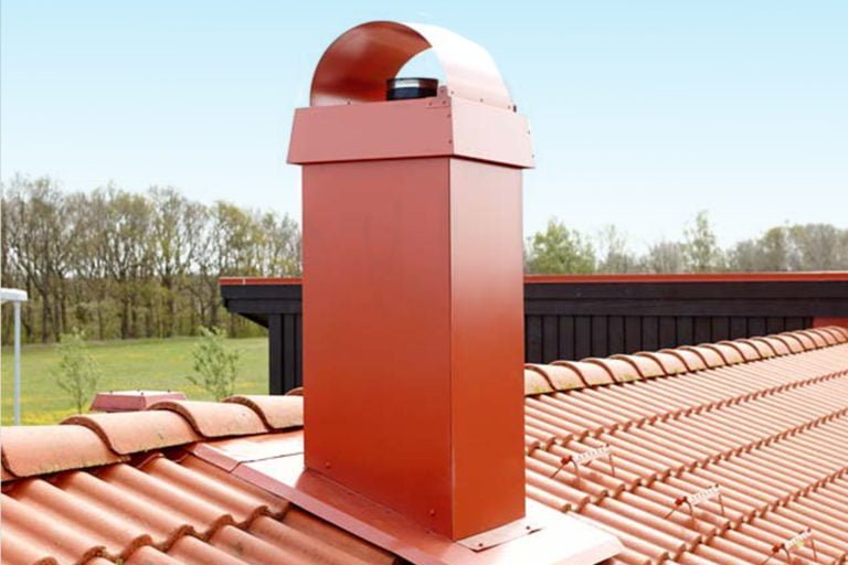 Rödfärgad skorsten med skyddande huv placerad på tak med tegelpannor, omgiven av grönskande landskap och klar himmel i bakgrunden.