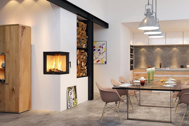 Modernt vardagsrum med en väggmonterad kamin, matbord, stolar och stilfull belysning. Vedförvaring och konstverk på väggen, träskåp på sidan.