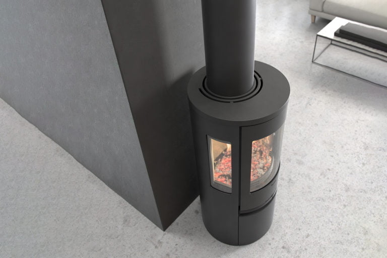 Svart, modern kamin placerad mot en grå vägg med glaspanel som visar glödande kol. Elegant inredningsdetalj i ett ljust, minimalistiskt rum.