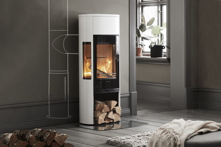 Modern vit kamin med brinnande eld och vedförvaring, placerad i ett stilrent vardagsrum med grå väggar och fönster med gröna växter.