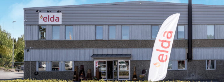 Företagsbyggnad med Elda-logotyp och flagga utanför. Grå fasad, stora fönster och grönska i förgrunden. Klarblå himmel. Nyckelord: Elda, företagsbyggnad.