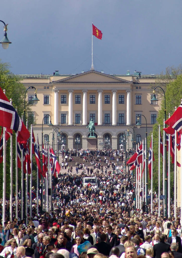 Folkmassa på Karl Johans gate i Oslo, Norge, med det kungliga slottet i bakgrunden och norska flaggor längs vägen.