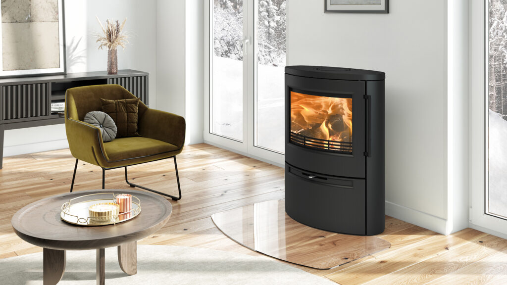 Modernt vardagsrum med braskamin, fåtölj och soffbord. Stora fönster visar snöig utsikt. Avslappnad atmosfär med ljus interiör och trädetaljer.