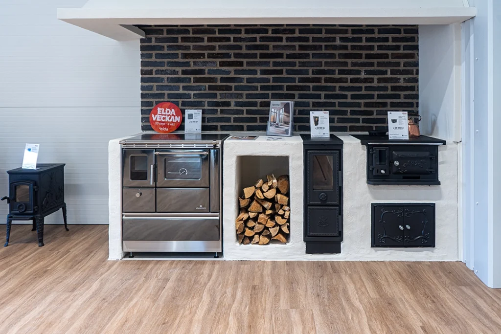Utställning av eldstad med vedspis, ved och informationsskyltar mot en mörk tegelvägg. Trägolv och ljusa väggar kompletterar interiören.
