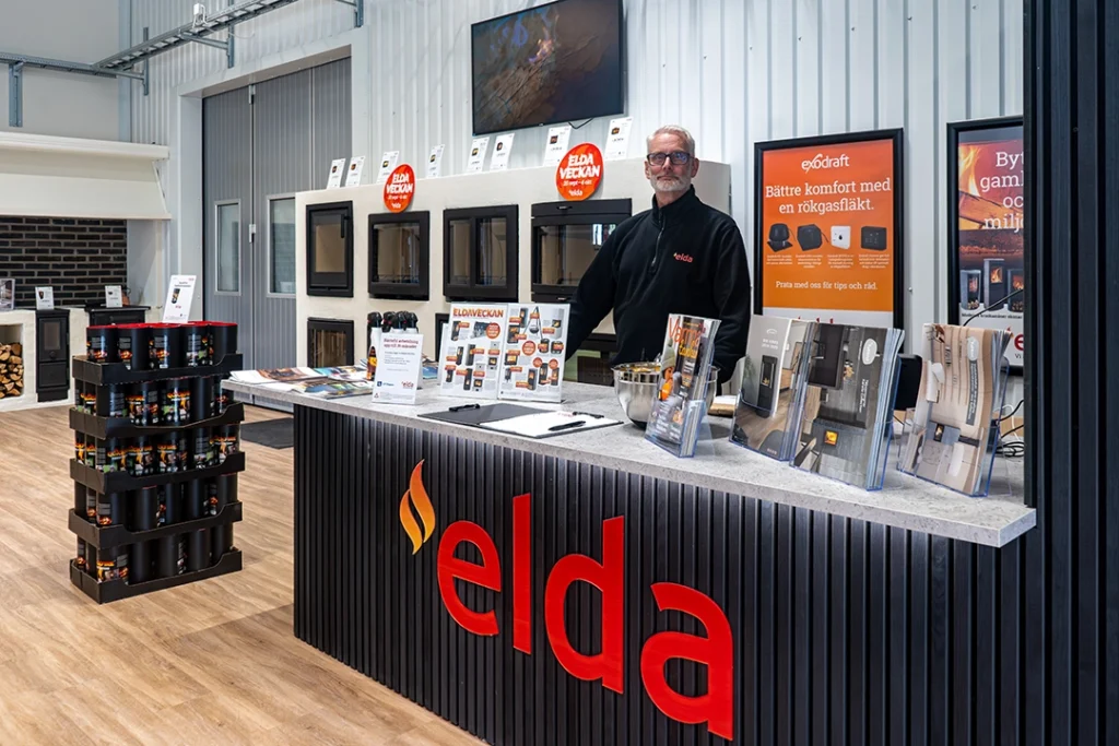 Utställningsrum i Eldabutiken med eldstäder, broschyrer och skyltar. Personal i svart tröja står bakom en disk märkt "Elda". Golvet är av trä.