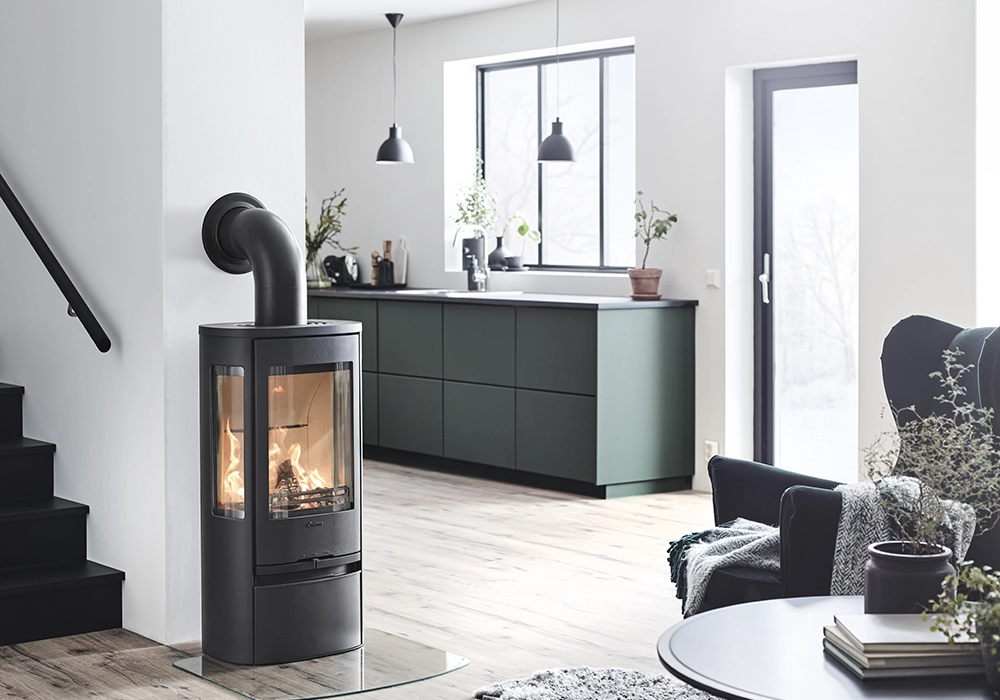 Modern kamin i ett stilrent vardagsrum med svart trappa, gröna köksskåp och stora fönster. Elden brinner inuti kaminen.