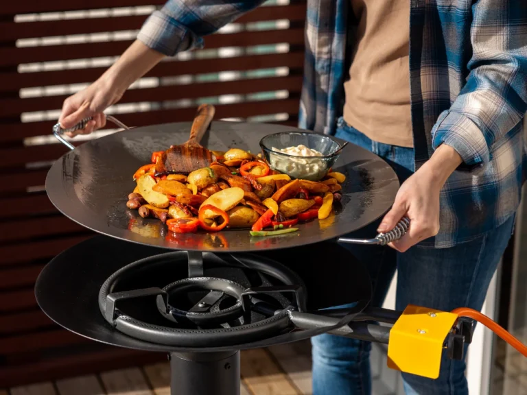 Person grillar grönsaker och potatis på en rund stekhäll utomhus. Detaljerad närbild av matlagning med stekspade och skål med sås.