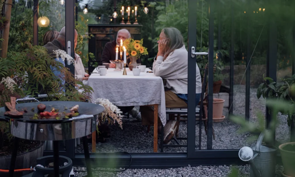 En grupp människor sitter runt ett dukat bord i ett grönskande växthus, omgivna av växter och levande ljus.