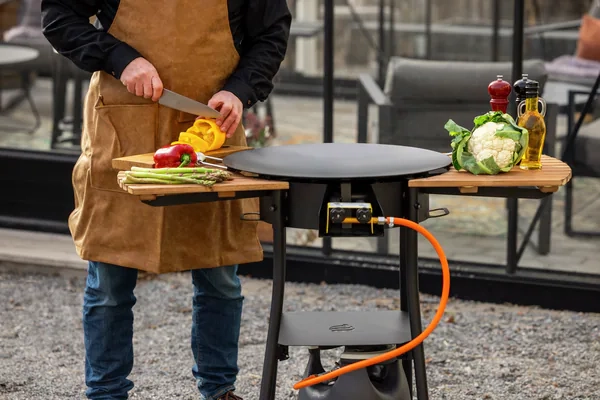 Person i förkläde skär paprika vid en utomhusgrill. Grönsaker och kryddor syns på sidoborden. Perfekt för grillning och matlagning utomhus.