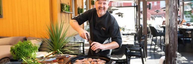 Kock i svart uniform lagar mat utomhus på en terrass, kryddar kött i en stor panna. Bakgrund med trävägg och grönska.
