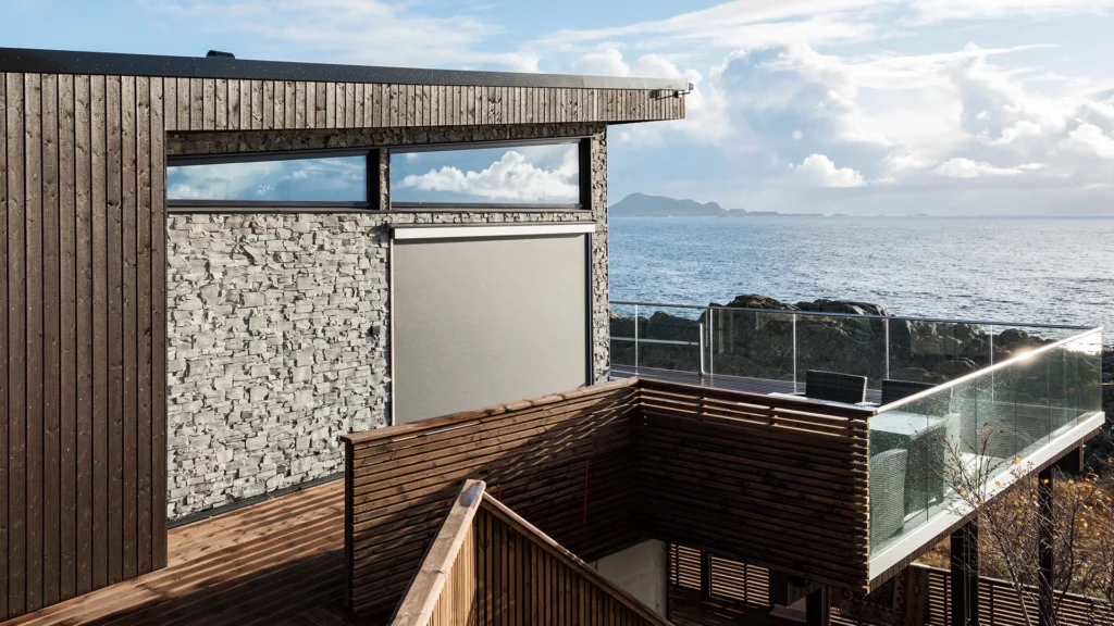 Modern villa vid havet med träfasad och glasräcke, omgiven av klippor och havsutsikt.