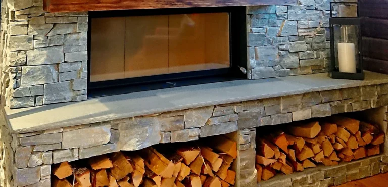 Modern eldstad med grå stenfasad, vedförvaring under och en svart lykta med vitt ljus på sidan. Perfekt för heminredning och värme.