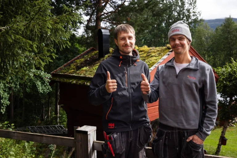 Två män i arbetskläder står utomhus framför en stuga med grönt tak. Den ena mannen har en Elda-mössa. Båda visar tummen upp. Skog i bakgrunden.