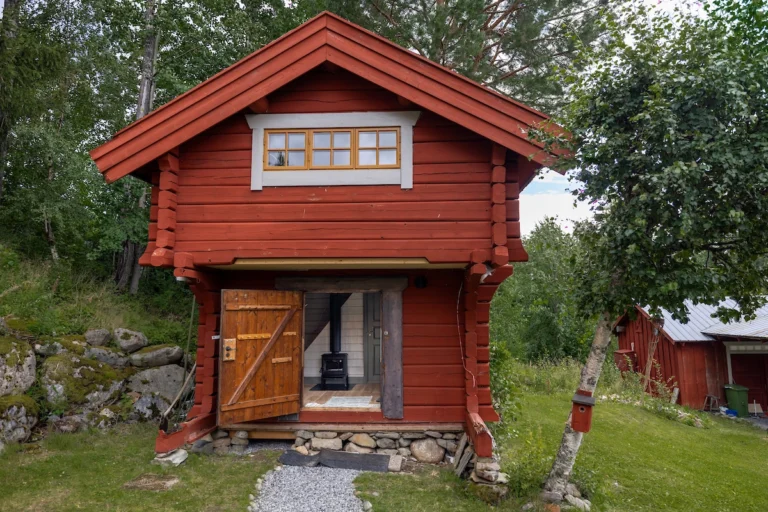 Rött timmerhus med öppen dörr, omgiven av grönskande träd och gräsmatta. En kamin syns inuti. Rustik och naturnära miljö.