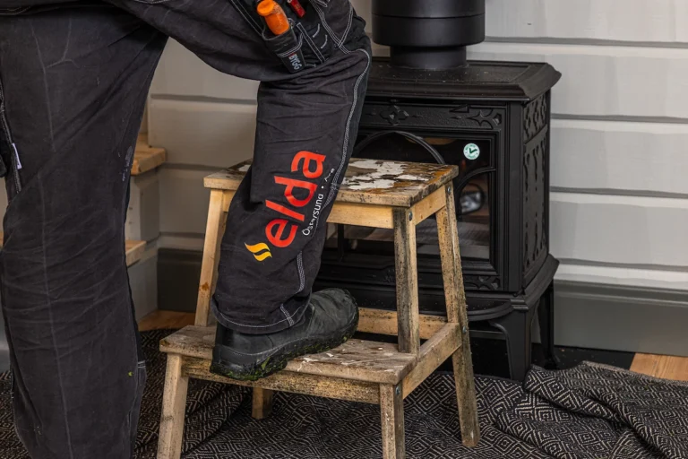 En person står på en träpall framför en svart kamin, klädd i arbetsskor och byxor med texten "elda".