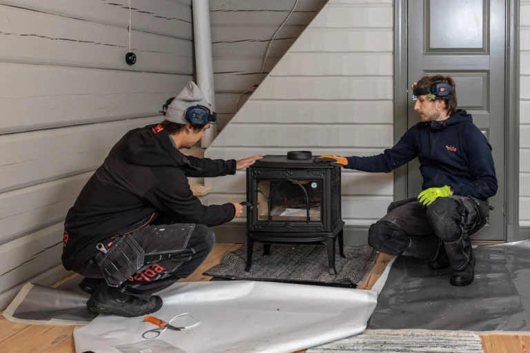 Två personer installerar en svart kamin i ett rum med träpanelväggar. De bär arbetskläder och skyddsutrustning. Verktyg och material syns på golvet.