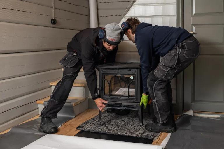 Två personer installerar en vedeldad kamin i ett rum med träväggar. De bär arbetskläder och hörselskydd.