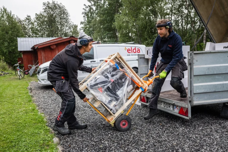 Två personer lastar av en vedkamin från ett släp med hjälp av en pirra. En bil med Elda-logotypen står i bakgrunden.