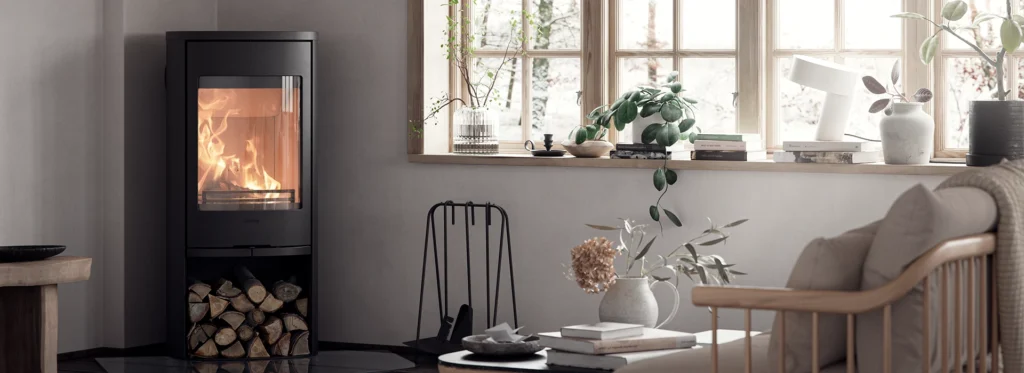 Modern braskamin i ett stilrent vardagsrum med vedförvaring under. Fönsterbräda med växter och böcker, ljus inredning och bekväm soffa i förgrunden.