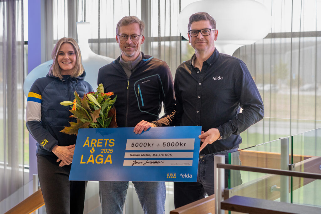 Tre personer poserar med en stor blå check och en blombukett inomhus, med texten "Årets Laga 2025" och summan 5000 kr.