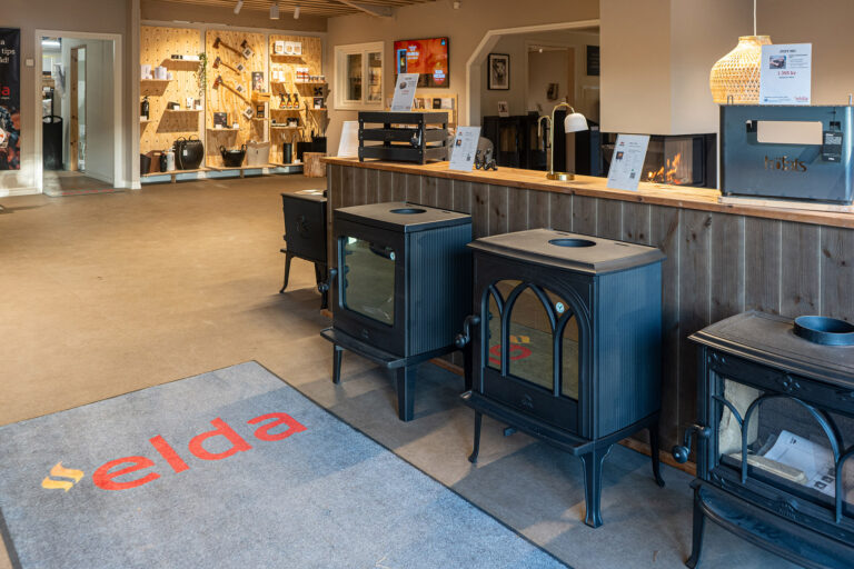 Utställning av eldstäder och kaminer i en butik, med olika modeller placerade på rad längs en träpanel. Eldabutiken-logga synlig på golvet.