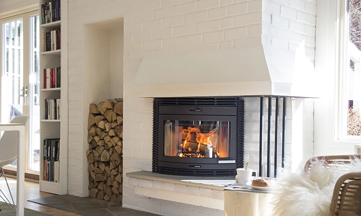 Vit tegelvägg med en modern braskamin i vitmålad omgivning. Elden brinner i kaminen. Ved staplad bredvid. Mysig inomhusmiljö med naturligt ljusinsläpp.