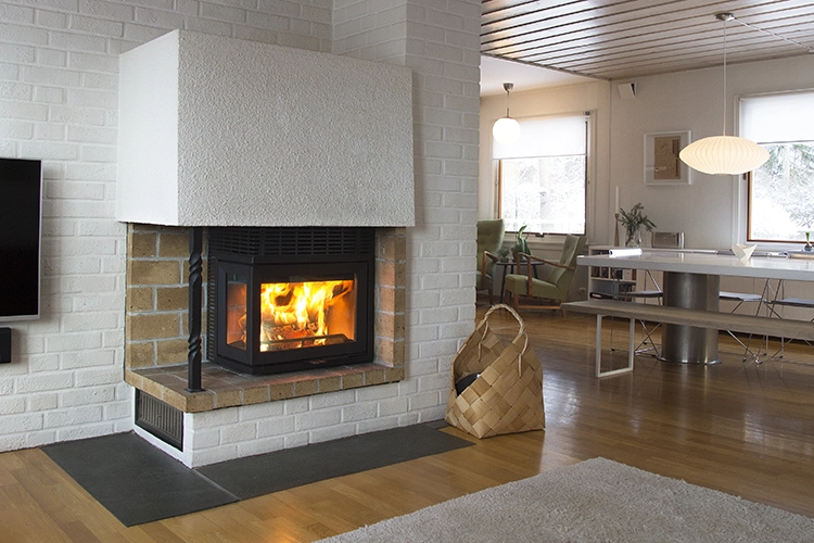 Modern braskamin i ett ljust vardagsrum med parkettgolv, vitmålade väggar och en flätad vedkorg bredvid. Elden brinner klart i kaminen.