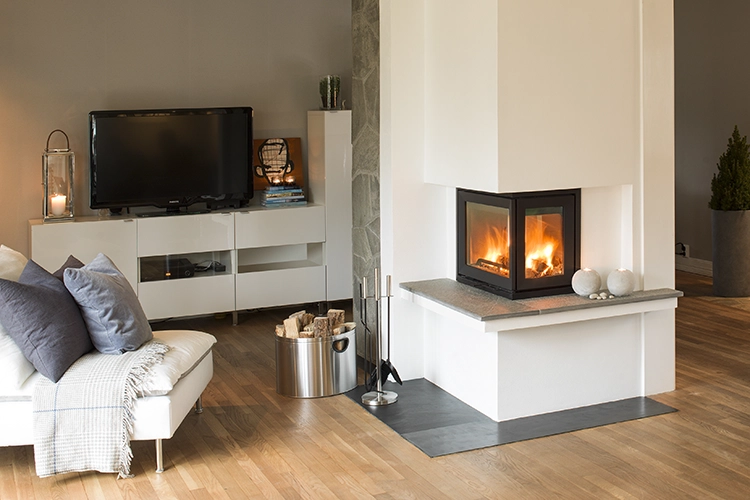Vardagsrum med modern hörnspis, tänd eld, vit inredning och trägolv. Soffa med kuddar och pläd, TV på vit skänk och vedkorg bredvid spisen.