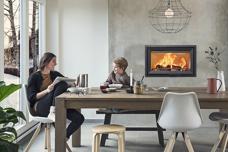 Två personer sitter vid ett matbord i ett modernt vardagsrum med en brinnande väggkamin, stora fönster och en hängande lampa.