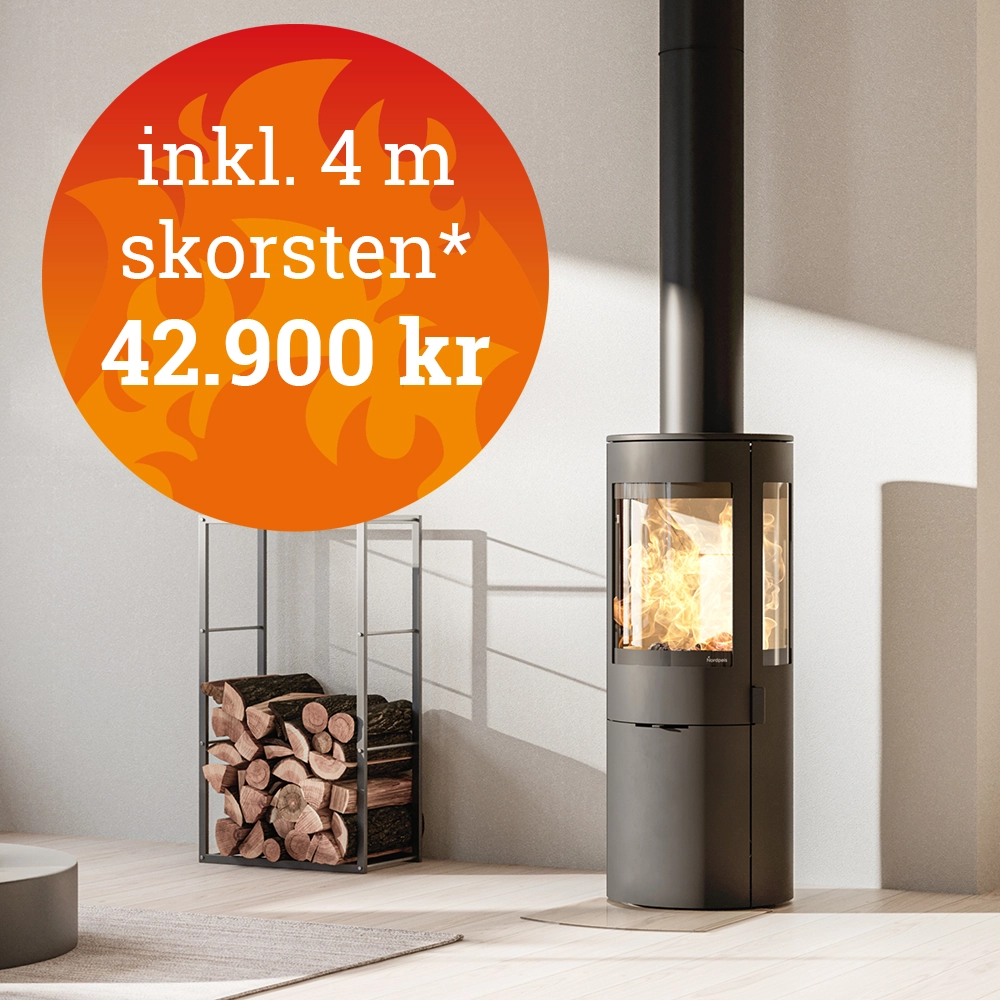 Modern vedkamin med glaslucka och brinnande eld, pris 42.900 kr inklusive 4 meter skorsten. Vedförvaring med staplade vedträn i bakgrunden.