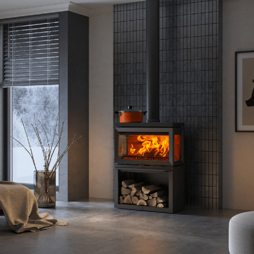 Modern vedeldad kamin med brinnande eld, omgiven av ved. En orange kastrull står på kaminen. Vinterlandskap syns genom fönstret i bakgrunden.