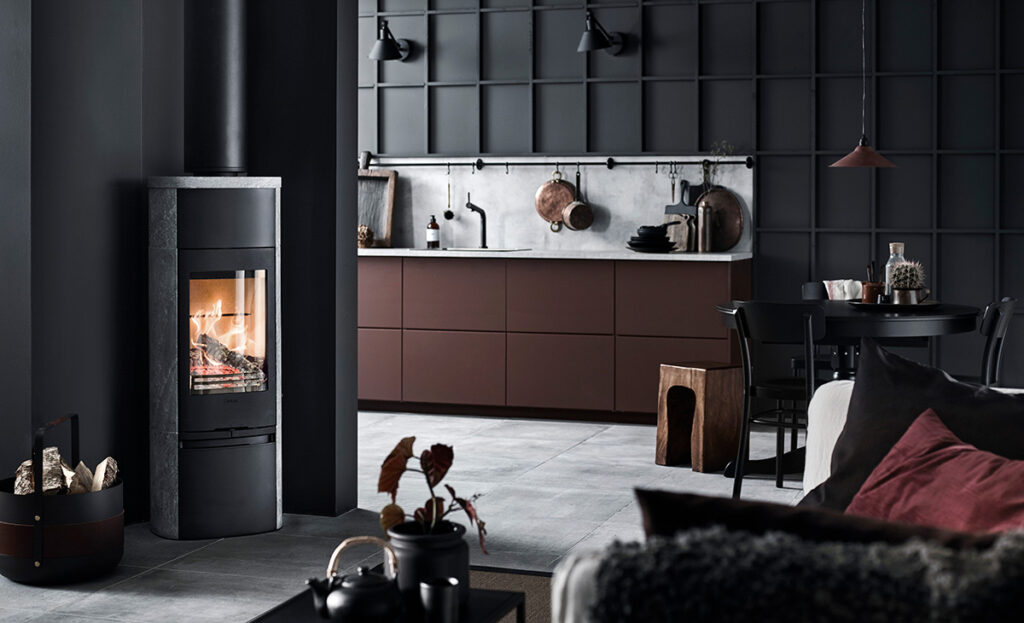 Modernt kök med mörka väggar, centralt placerad vedeldad kamin och stilfull inredning. En blandning av industriell och skandinavisk design skapar en varm, inbjudande atmosfär.