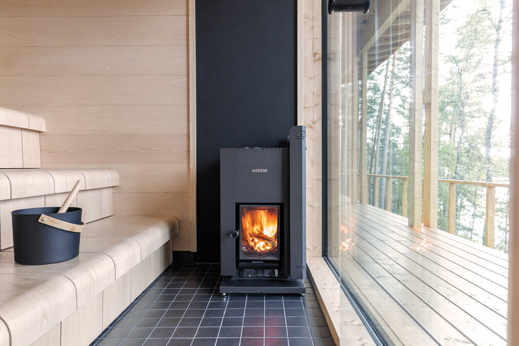 Modern bastu med vedeldad kamin, träbänkar och stora glasfönster med utsikt över skogslandskap. En svart hink med skopa står på golvet.