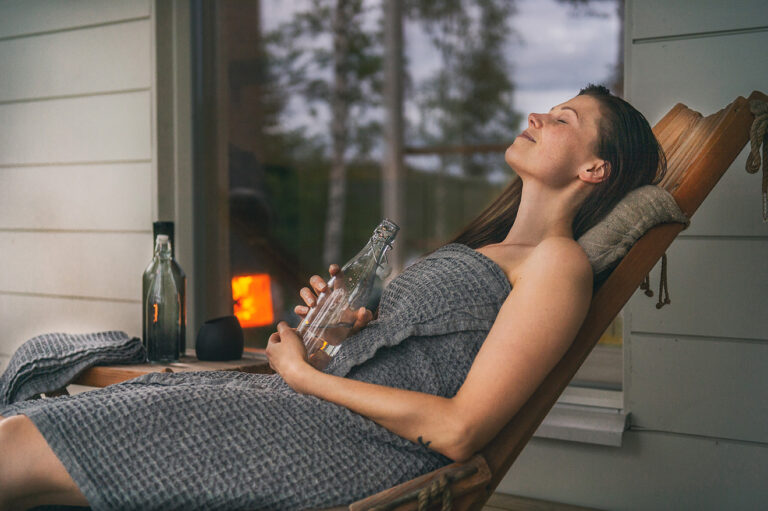 Kvinna i grå handduk slappnar av i solstol med glasflaska. Bakgrund med fönster och brinnande ljus.