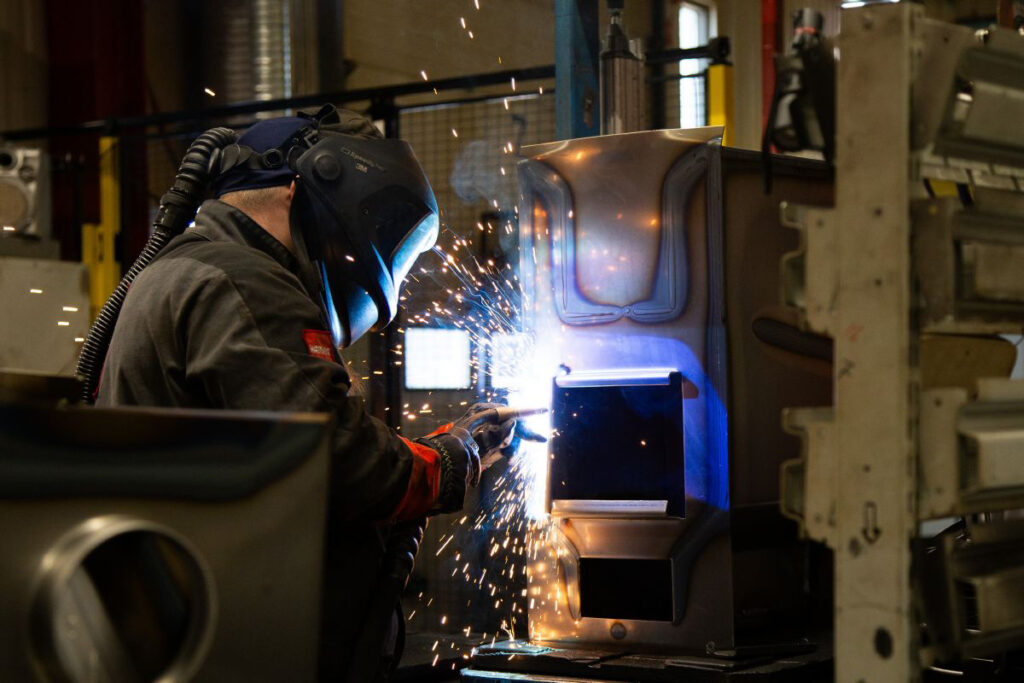 Person i skyddsutrustning svetsar metall i en industrimiljö, gnistor flyger. Fokus på hantverk och säkerhet i metallbearbetning.