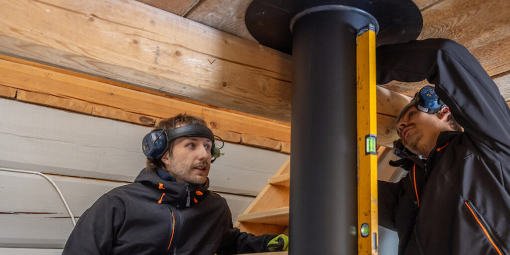 Två personer i arbetskläder monterar en kamin i ett trähus. De använder hörselskydd och en vattenpass för att säkerställa korrekt installation.
