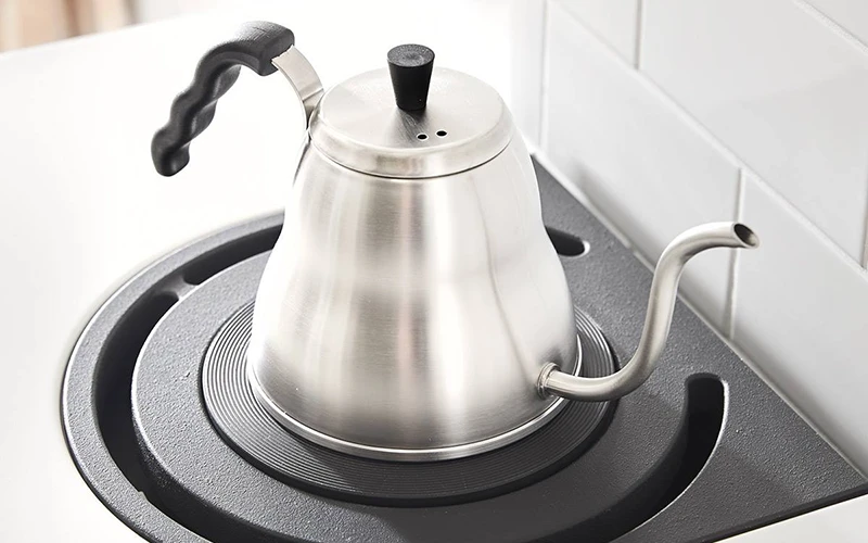 Rostfri vattenkokare med svanhals och svart handtag på en rund kokplatta i vitt kök.