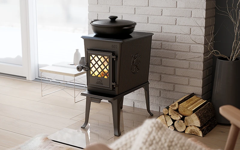 En svart vedeldad kamin står framför en vit tegelvägg. Ved ligger bredvid och en kastrull står ovanpå kaminen. Elden brinner inuti.