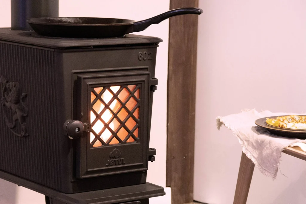 Vedeldad gjutjärnskamin från Jøtul med glaspanel, glödande eld inuti. En stekpanna ovanpå kaminen och en tallrik på ett bord bredvid.