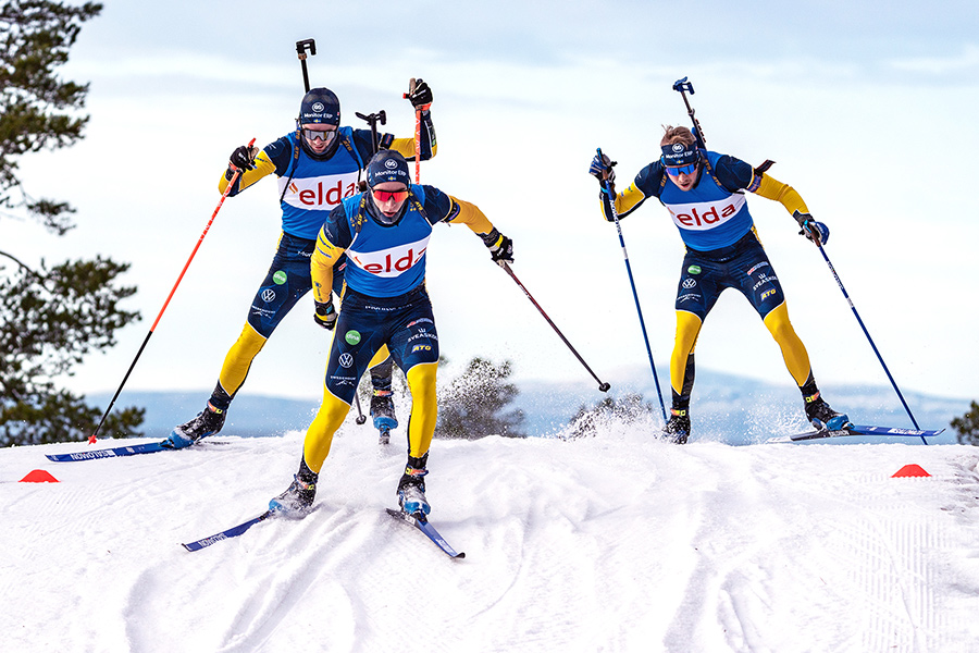 Tre skidåkare klädda i blå och gula dräkter deltar i en skidskyttetävling på snötäckt landskap, med gevär på ryggen och stavar i händerna.