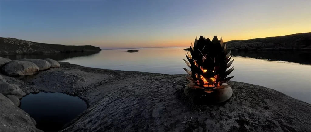 Bilden visar en eldkorg formad som en kotte, placerad på klippor vid vattnet i solnedgången. Elden lyser varmt mot den mörknande himlen.