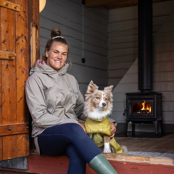 Kvinna i beige jacka och hund i gul regnrock vid vedspis i trästuga. Eldstad i bakgrunden.