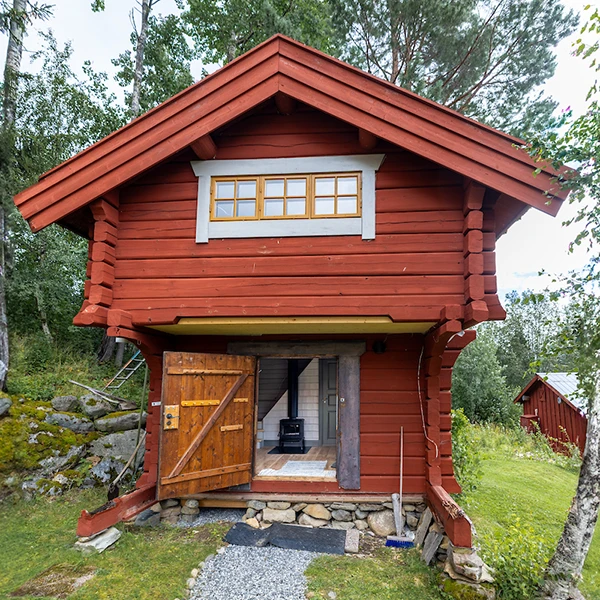Röd timmerstuga med öppen dörr, omgiven av grönska och träd. Stugan står på en stenfot och har en liten stig fram till ingången.