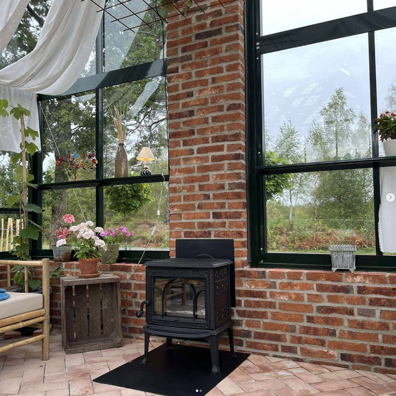 Braskamin i ett växthus med tegelväggar, omgiven av växter och blommor, skapar en mysig atmosfär.