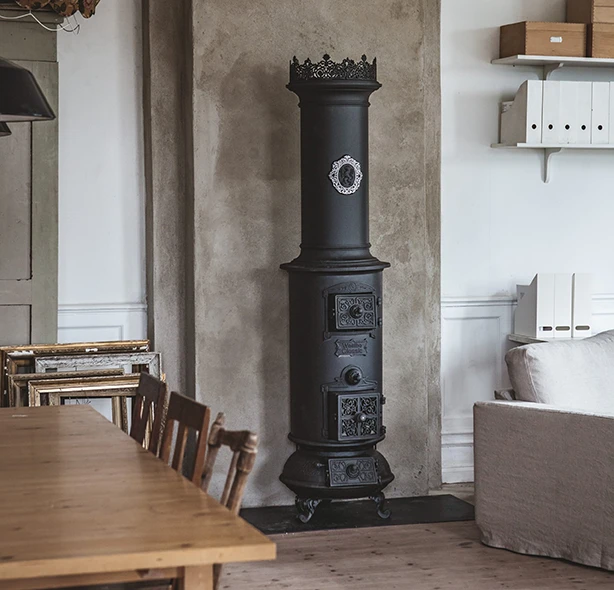 Svart gjutjärnskamin mot betongvägg i vardagsrum med träbord och hyllor. Kaminens detaljerade design ger en rustik känsla.
