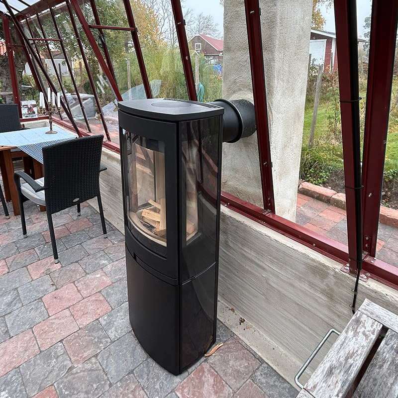 Modern kamin installerad i inglasat uterum med stenlagd golvyta, omgiven av trädgårdsmiljö. Nyckelord: kamin, uterum.
